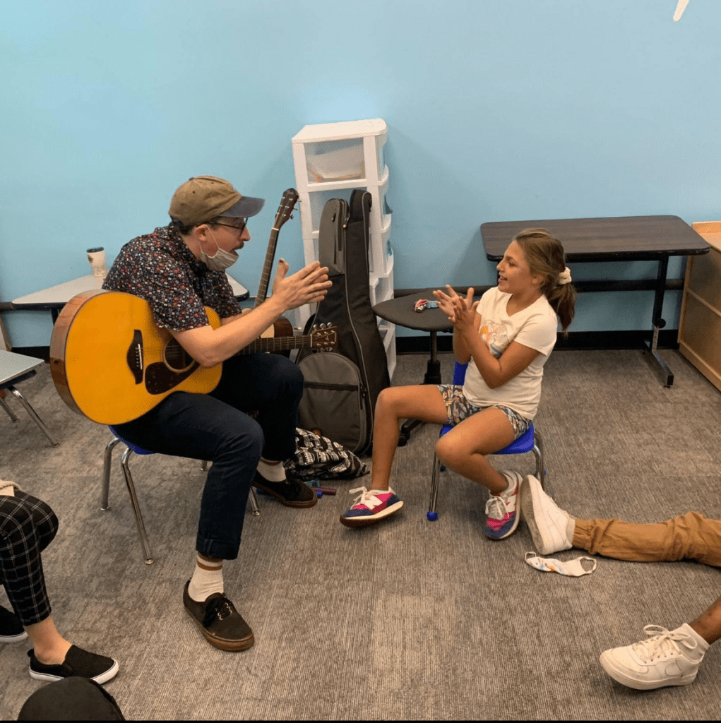 A photo of Maximo playing guitar and clapping in rhythm with a young child in a music therapy session. 