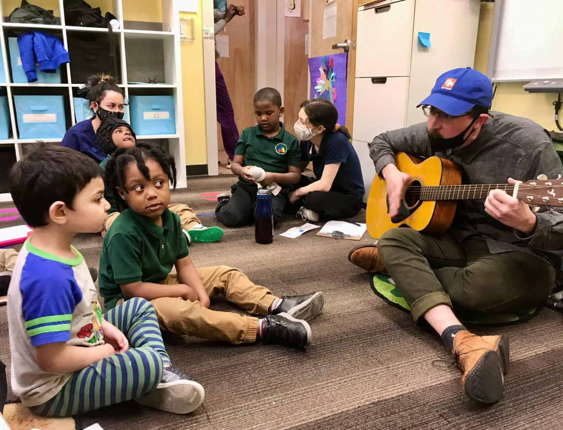 A photo of Maximo playing guitar in a music therapy session working with young children. 